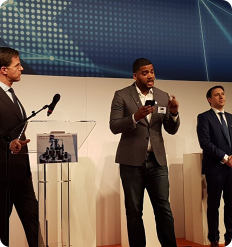 Three men in suits standing on stage, one speaking and holding a smartphone, with a microphone and a podium nearby.