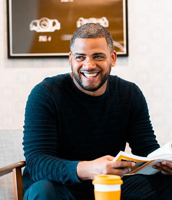 Smiling man with short hair and beard sitting indoors, holding a book and a yellow coffee cup on the table.