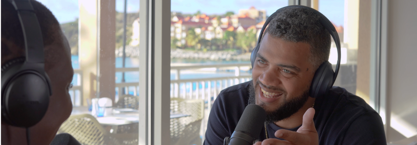 Two men wearing headphones talking into microphones with a waterfront view through a window behind them.