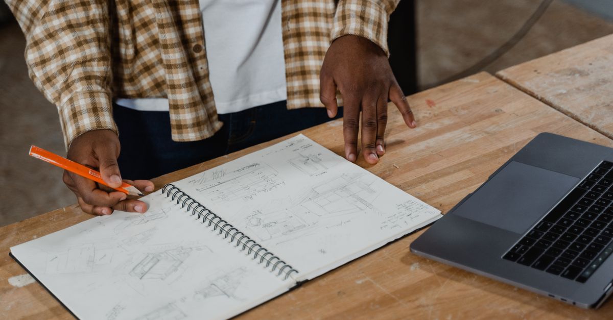 A craftsman examines detailed workshop plans, sketchbook, and laptop on the workbench.