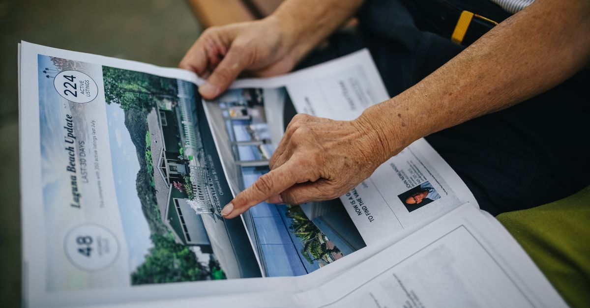 A senior adult's hands holding and reading a newspaper while seated on an outdoor bench.