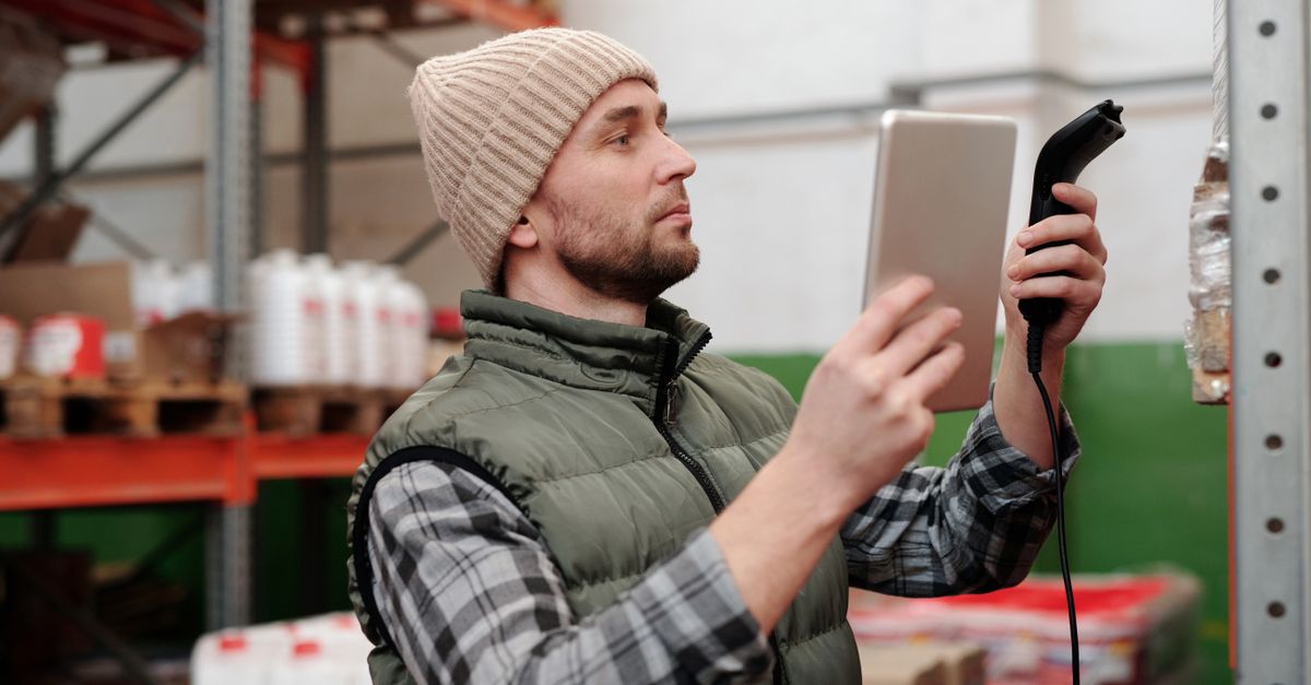 Man in beanie and vest using a scanner in a warehouse for inventory control.