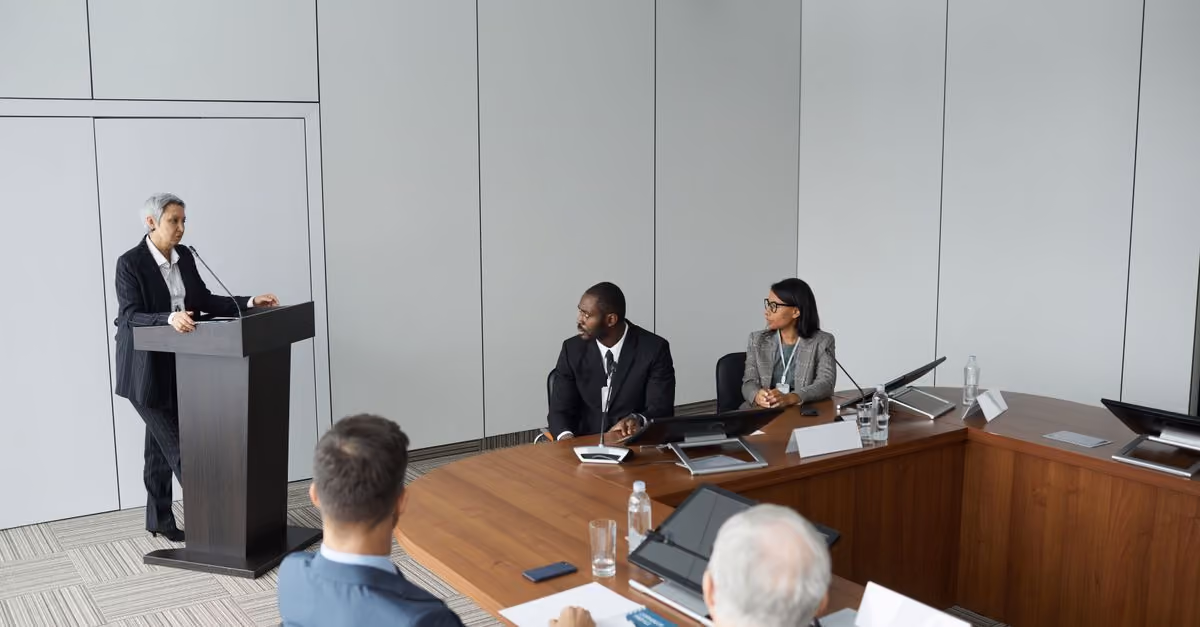 Diverse group of professionals in a formal meeting setting with a speaker at the podium.