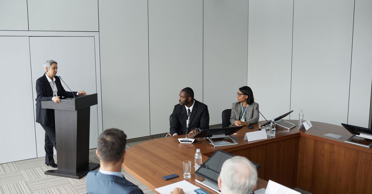Diverse group of professionals in a formal meeting setting with a speaker at the podium.