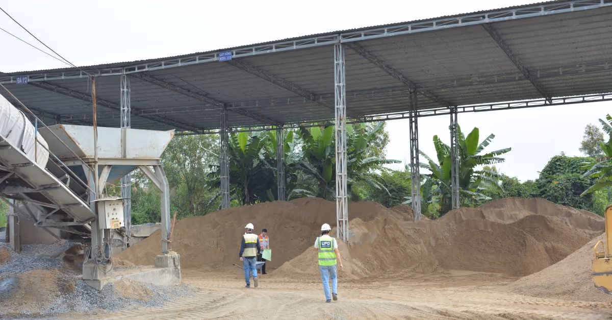 Industrial construction site with workers inspecting sand piles under metal roofing.