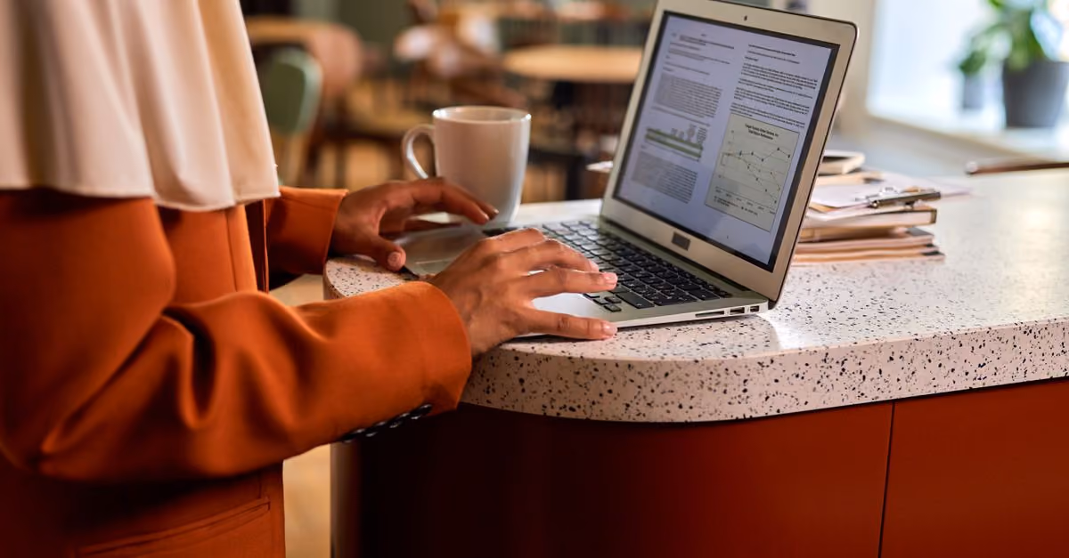 Adult woman typing on a laptop at a modern office counter, coffee beside her in a casual indoor setting.