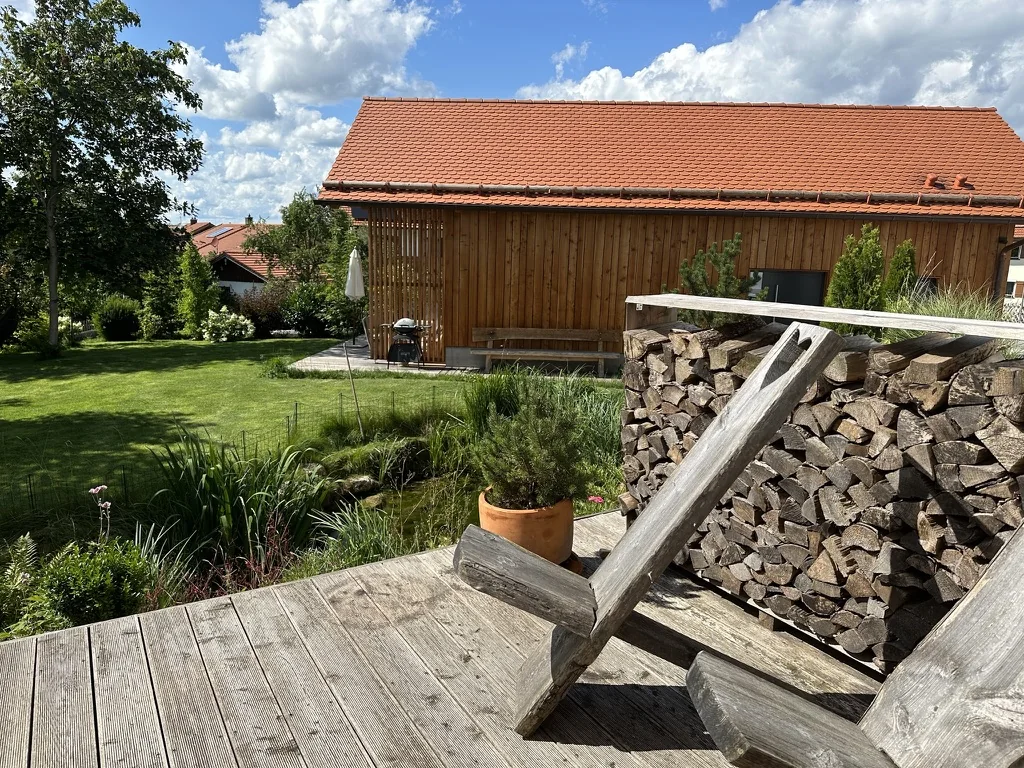 Holzterrasse mit Bergstuhl und Kaminholz, Blick auf Garten, Teich und Holzchalet