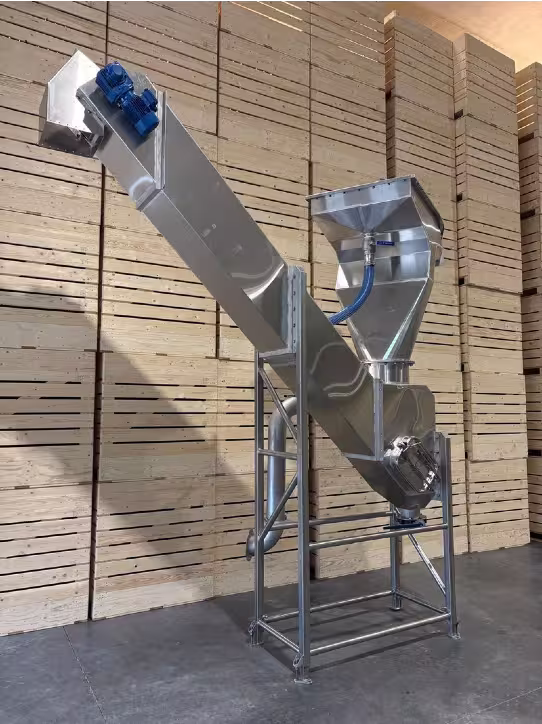 Stainless steel inclined conveyor machine with hopper and motor, positioned inside a warehouse lined with wooden storage crates.