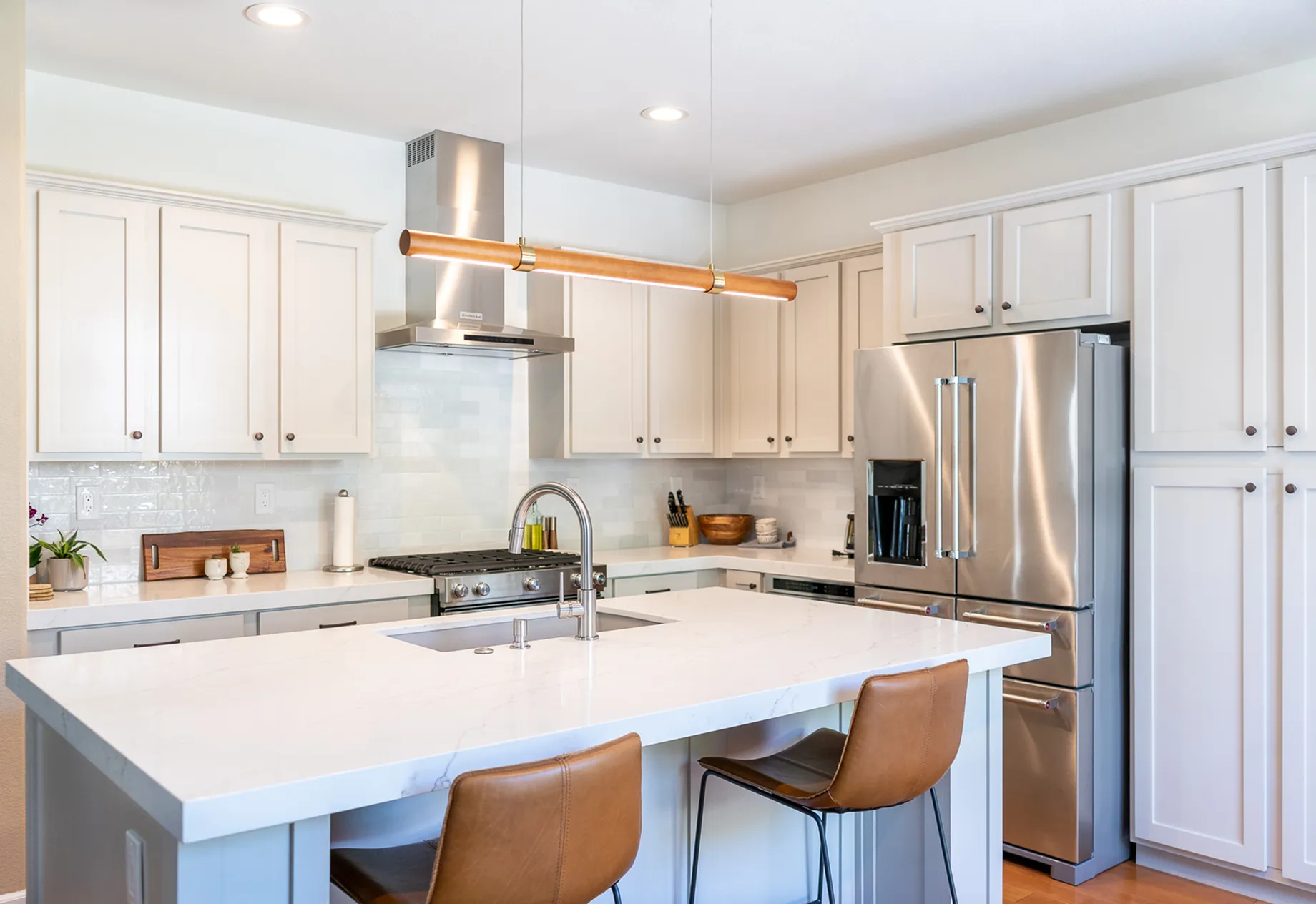 Modern kitchen with white cabinets, marble countertop island with sink, two brown leather chairs, stainless steel refrigerator and stove.