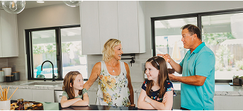 Family of four smiling and talking together in a bright kitchen with large windows.
