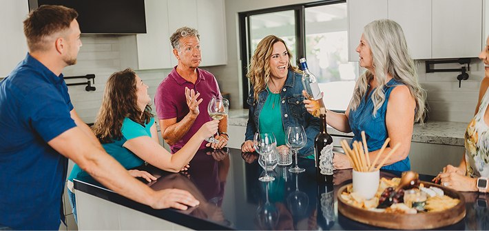 Group of six adults socializing around a kitchen island with wine glasses and a charcuterie board.