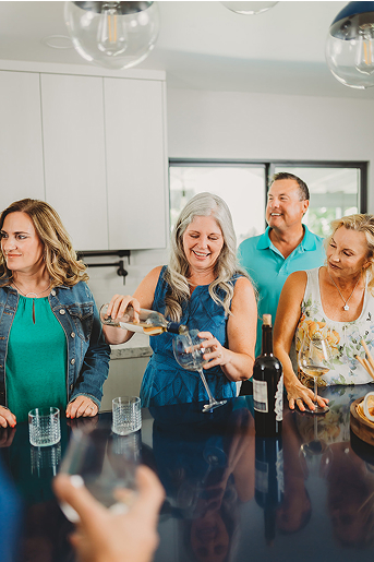 Group of four adults smiling and pouring wine while standing around a kitchen island.