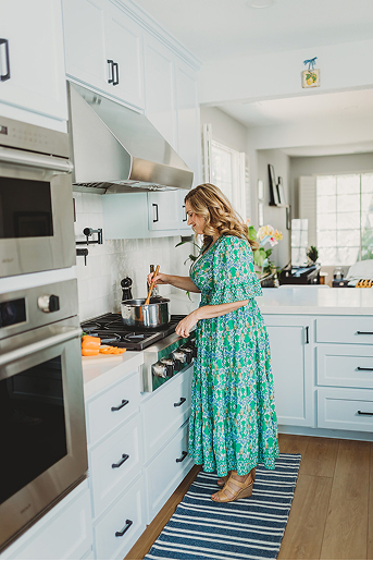 Woman in green floral dress cooking with a pot on a stove in a bright, modern kitchen.