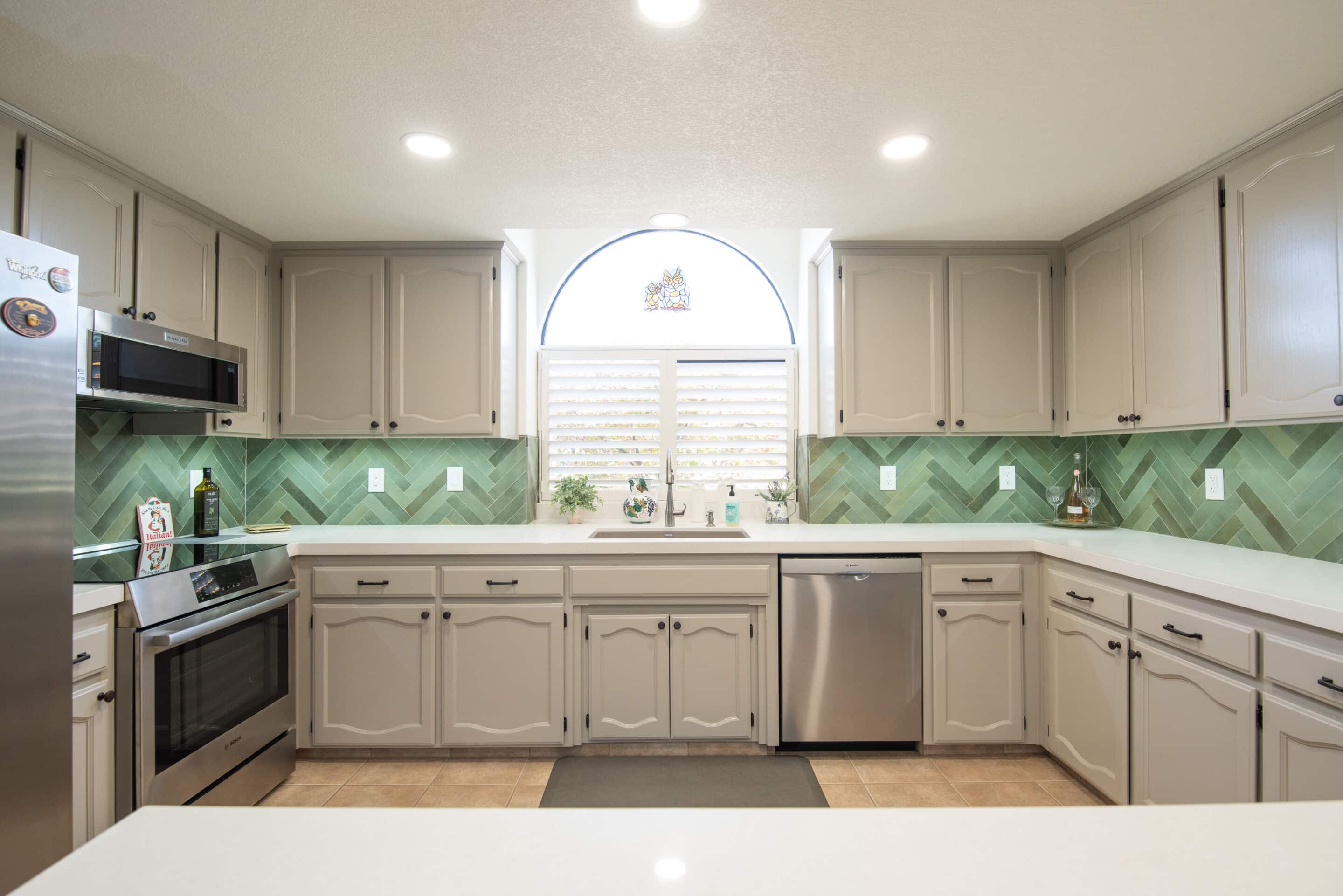 Bright kitchen with beige cabinets, green herringbone tile backsplash, stainless steel appliances, and an arched window above the sink.