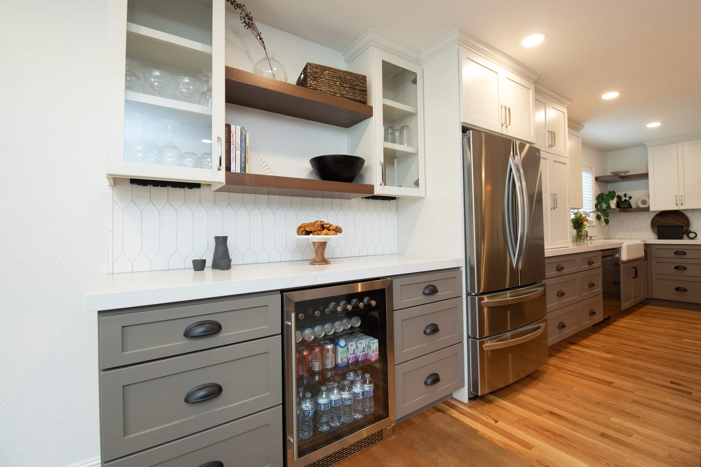 Modern kitchen with gray lower cabinets, white upper cabinets, a stainless steel refrigerator, a built-in beverage cooler, and wooden shelves with decor and croissants on a cake stand.