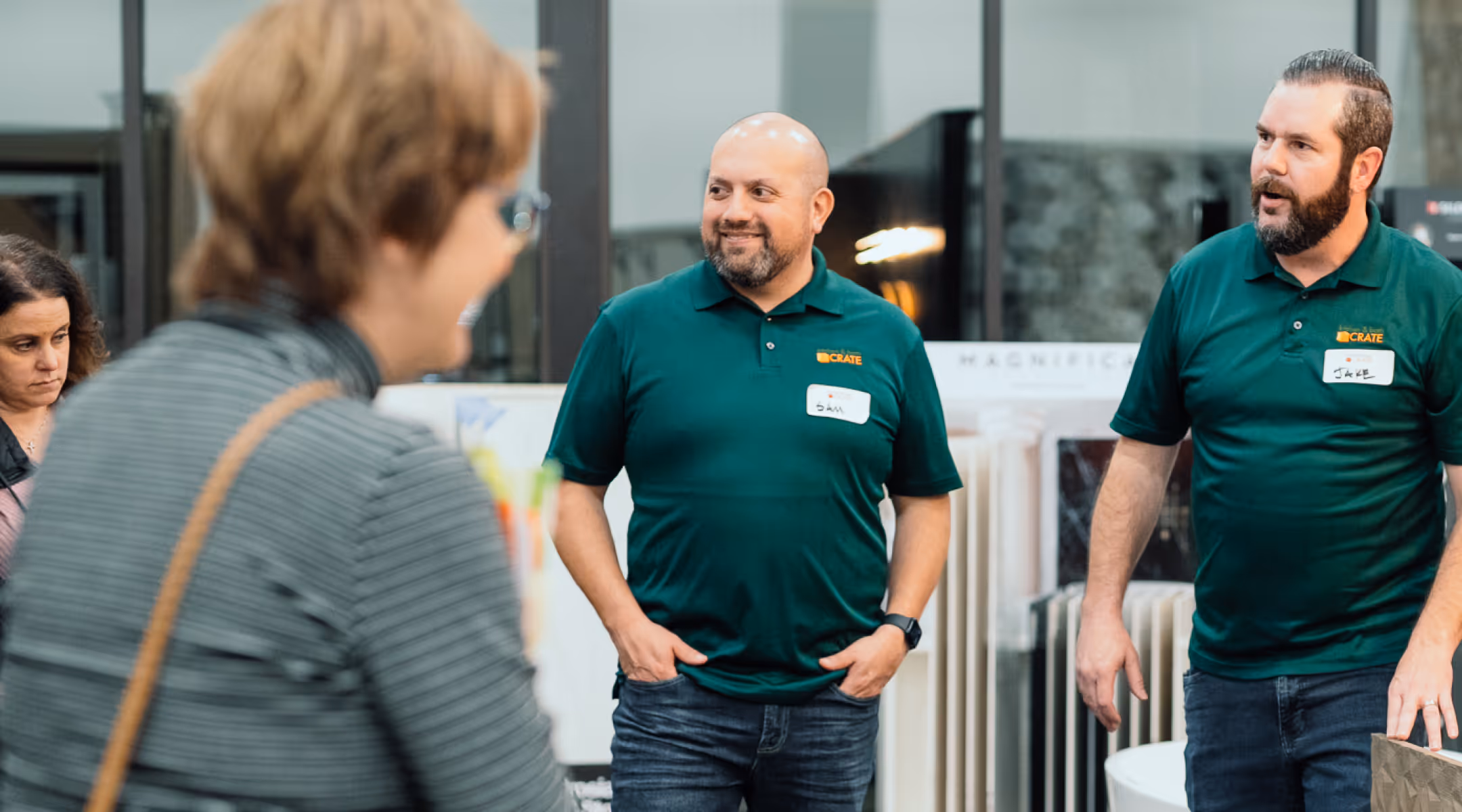 Two men in green polo shirts with name tags talking at an indoor event while two women listen nearby.