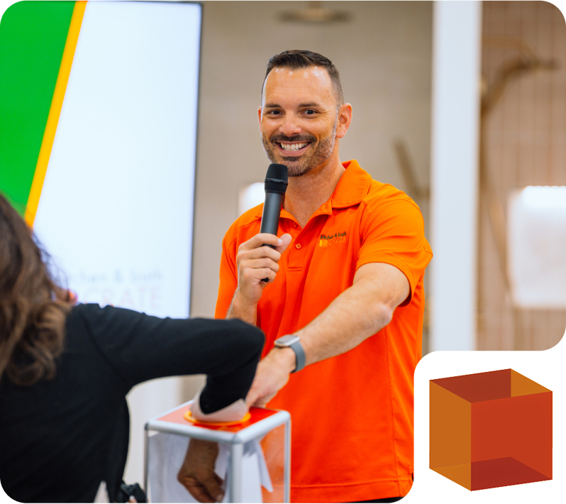Man in an orange polo shirt speaking into a microphone while helping a person press a large red button.