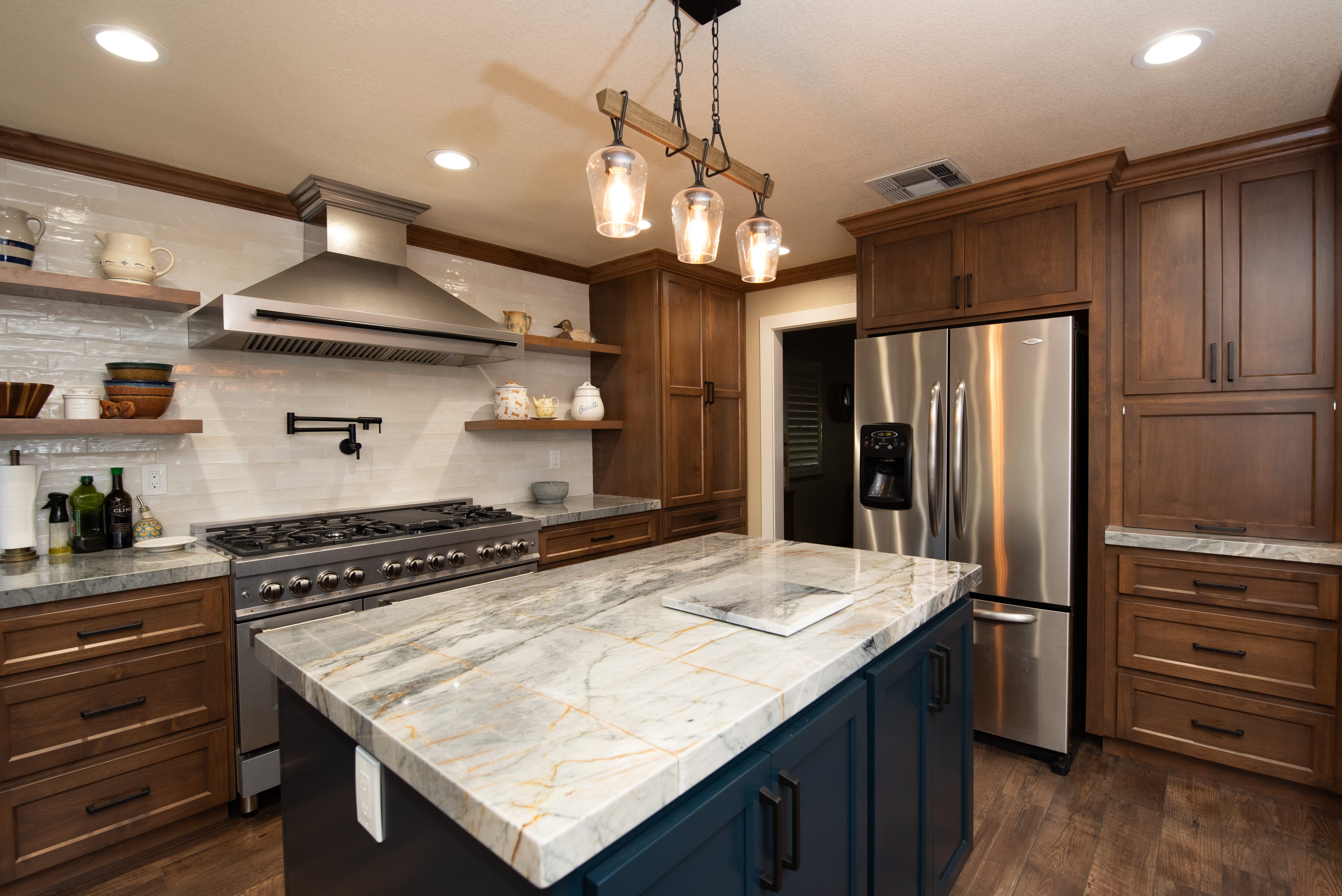Modern kitchen with marble island countertop, stainless steel refrigerator, gas stove, wooden cabinets, and hanging pendant lights.
