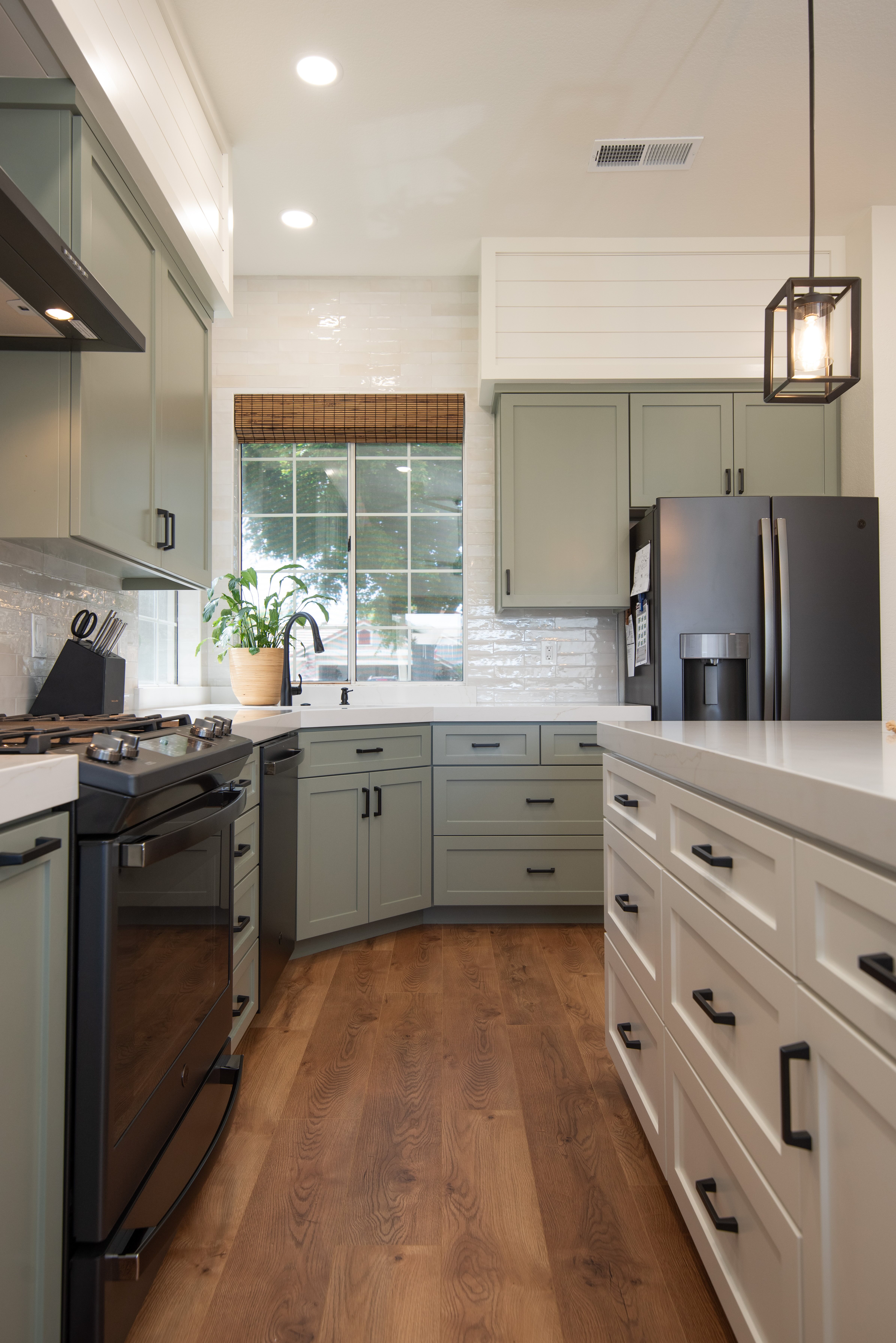 Modern kitchen with light green cabinets, white countertops, wooden floor, black appliances, and a window with a bamboo shade.