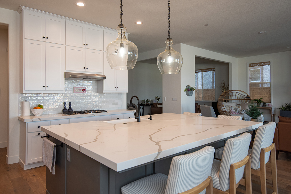 Modern kitchen with large marble island, white cabinetry, two glass pendant lights, and four beige striped chairs.
