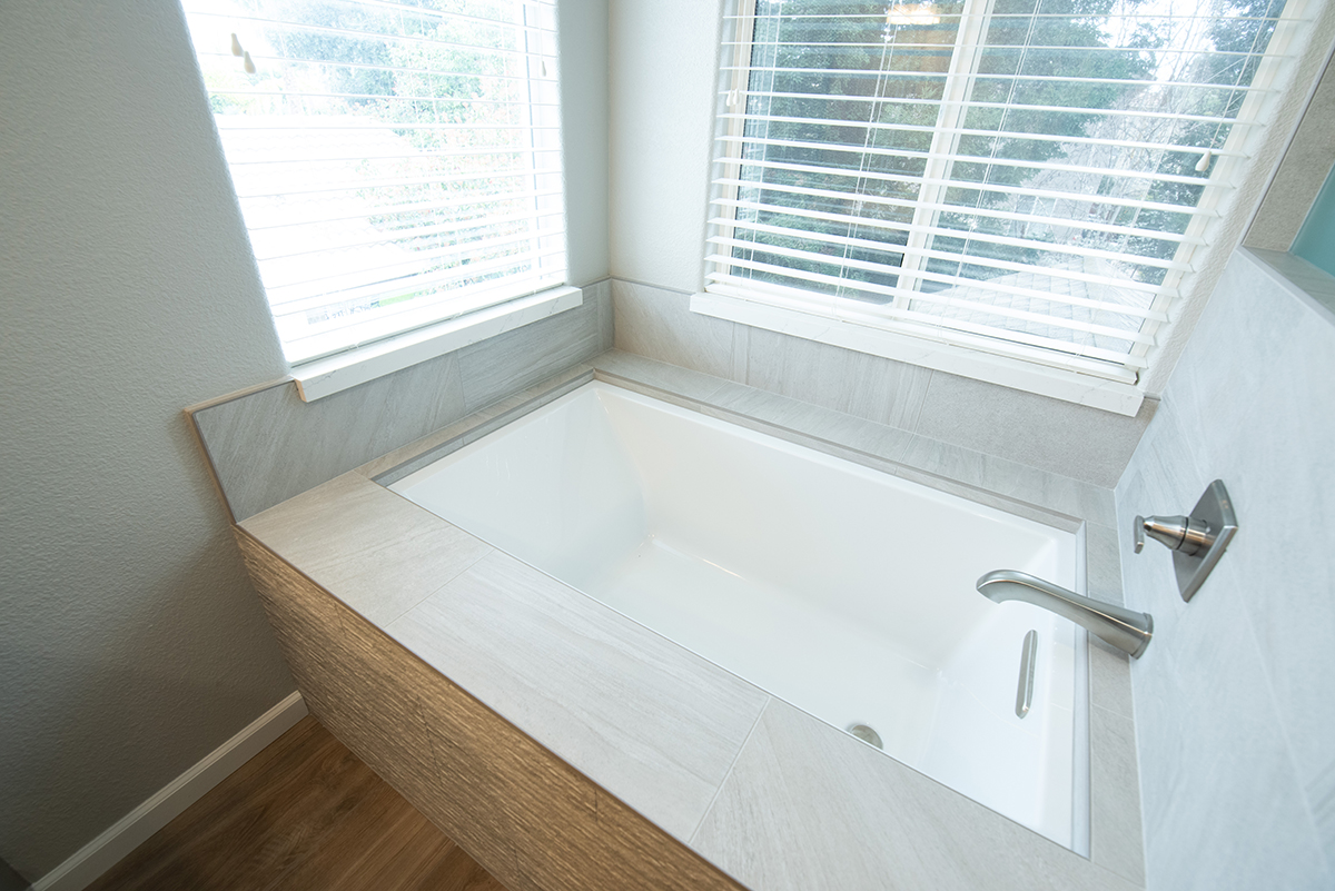 Modern rectangular white bathtub with gray tile surround and silver faucet, situated under two windows with white blinds.