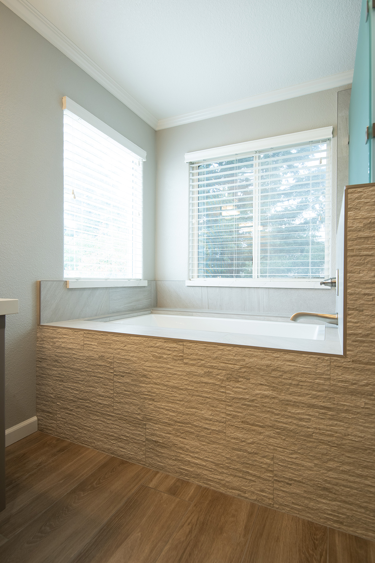 Modern bathroom with a large built-in bathtub surrounded by textured beige tiles and two windows with white blinds.