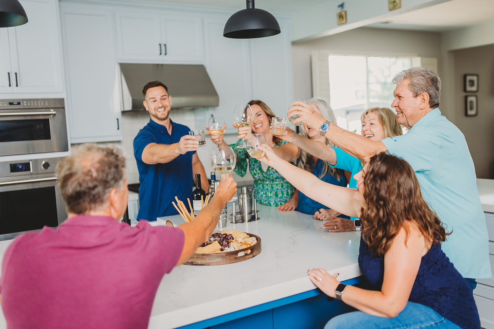 Group of six adults gathered around a kitchen island toasting with drinks, smiling and enjoying a social moment.