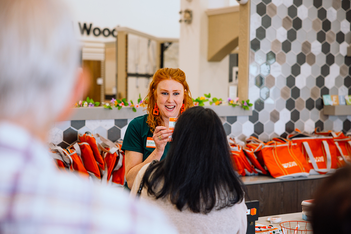 A woman with red hair and green shirt holds a product while talking to another person at an event with orange bags in the background.