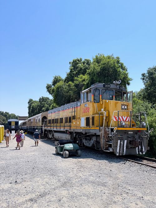 Yellow and gray diesel locomotive train numbered 2658 parked on tracks with people walking nearby on a gravel path under a clear blue sky.