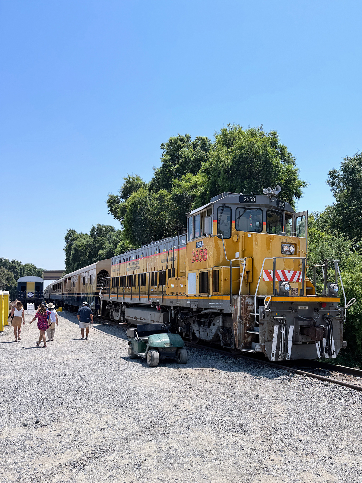 Yellow and gray diesel locomotive train numbered 2658 parked on tracks with people walking nearby on a gravel path under a clear blue sky.