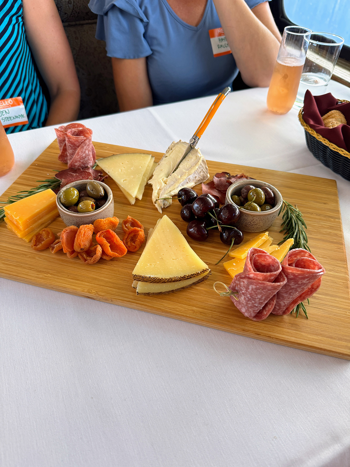 Charcuterie board with various cheeses, dried apricots, green and black olives, cherries, rolled salami, and rosemary sprigs on a wooden board.
