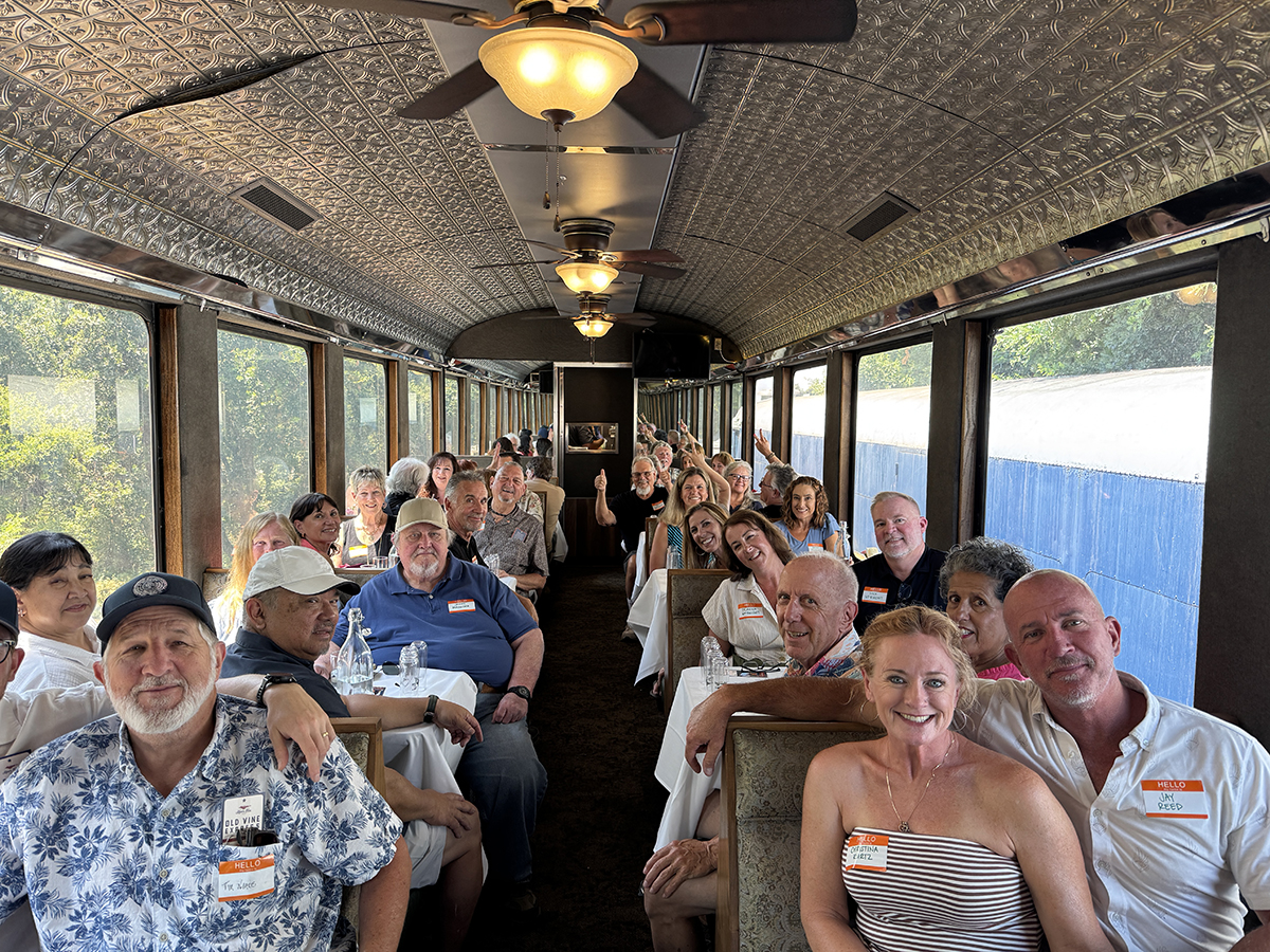 Group of people seated at tables inside a vintage-style train car with large windows and ornate ceiling.