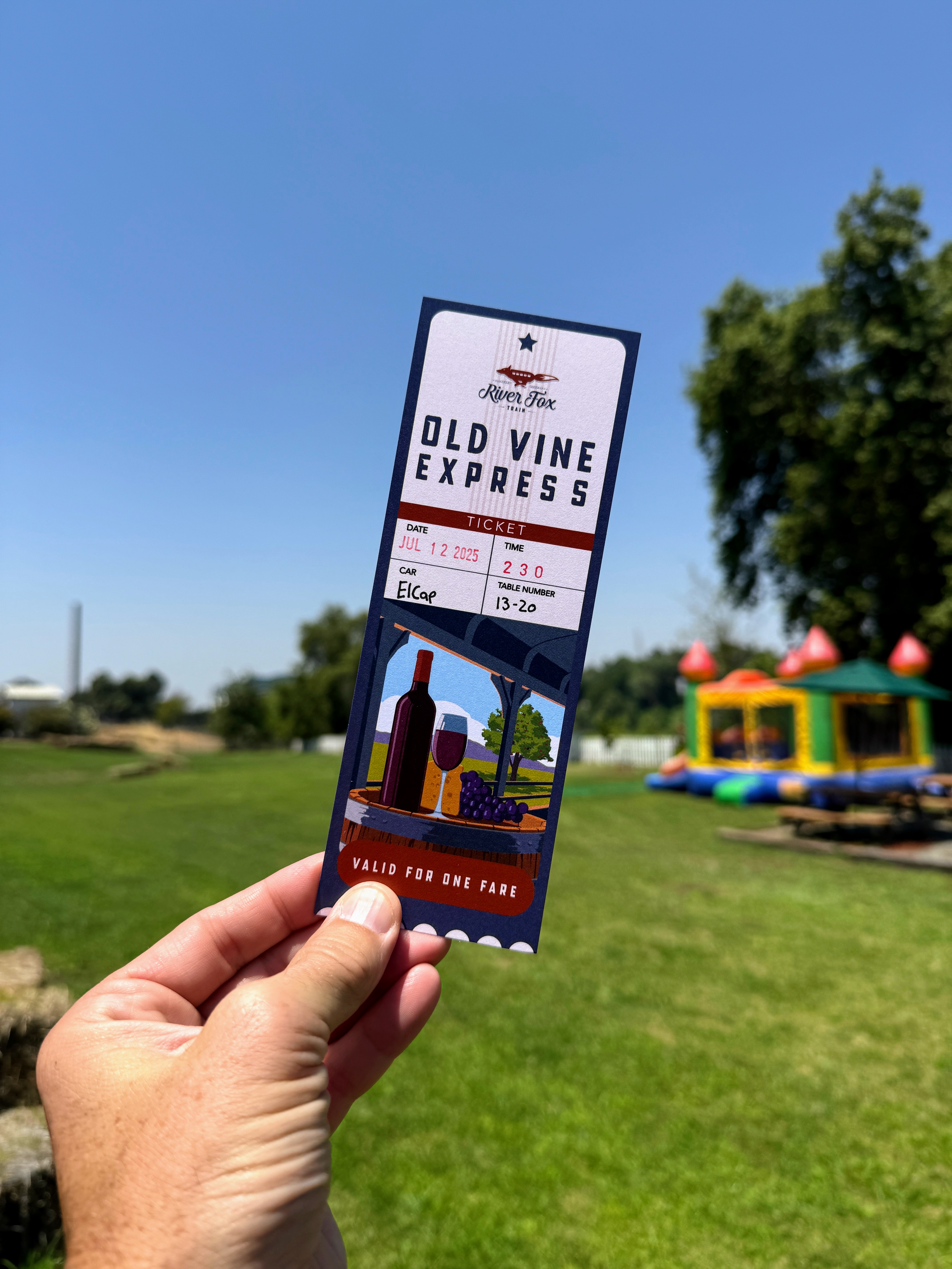 Hand holding a River Fox Train Old Vine Express ticket with a picnic scene and green park in the background.