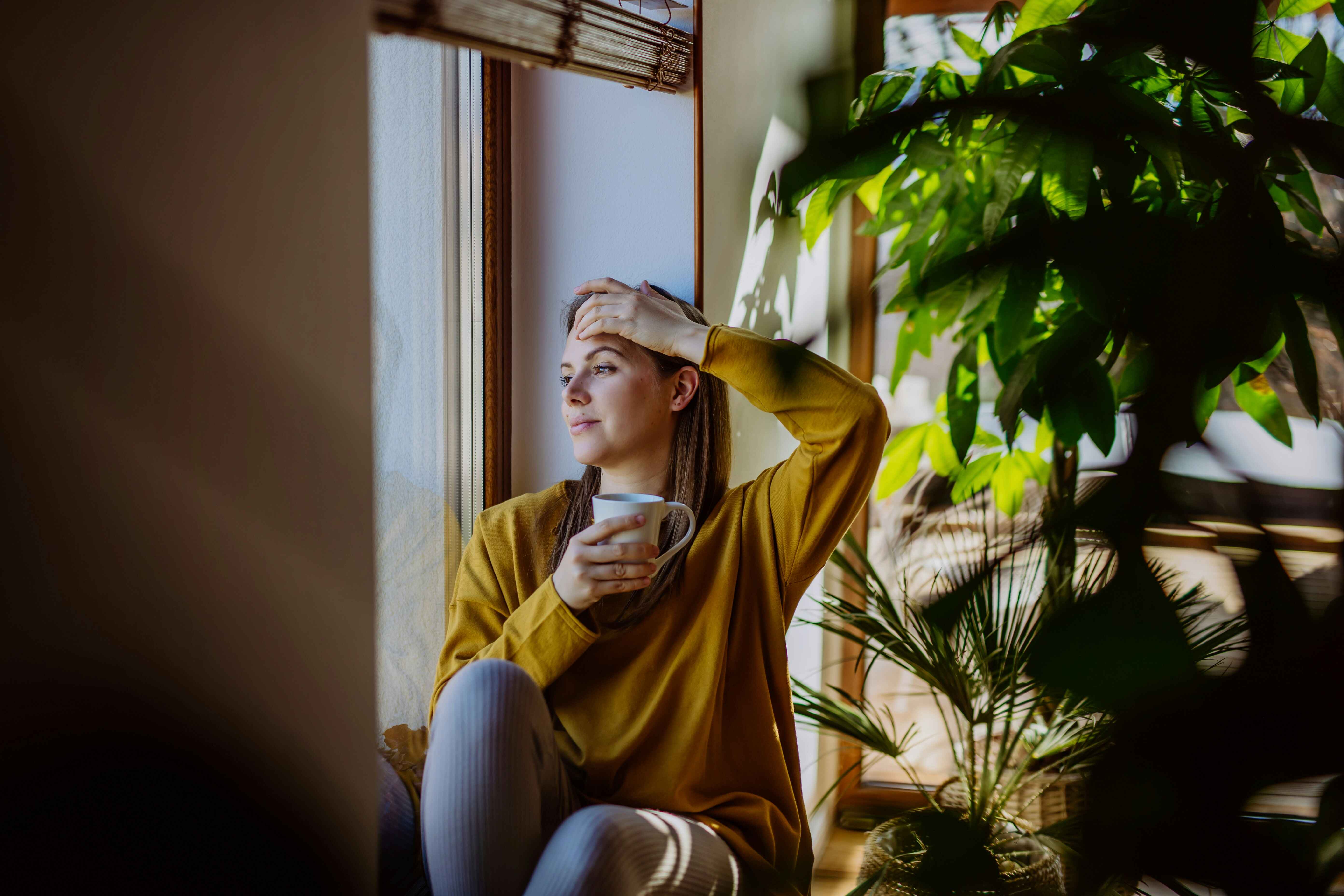Woman in a yellow sweater holding a mug and looking out a window, reflecting during therapy