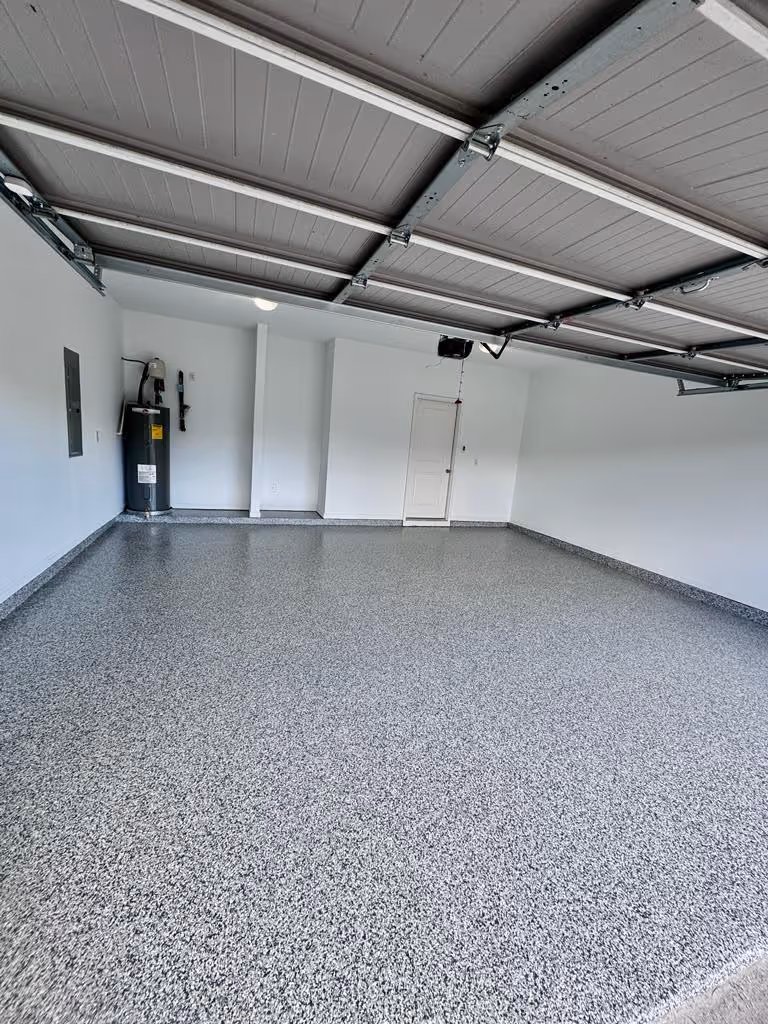 Empty garage with newly coated speckled gray epoxy floor, white walls, closed gray ceiling garage door, water heater, and a white door on the back wall.