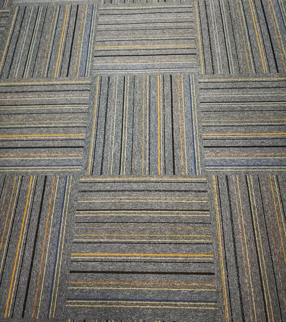 Close-up of carpet tiles featuring alternating vertical and horizontal striped patterns in gray, yellow, black, and blue tones.
