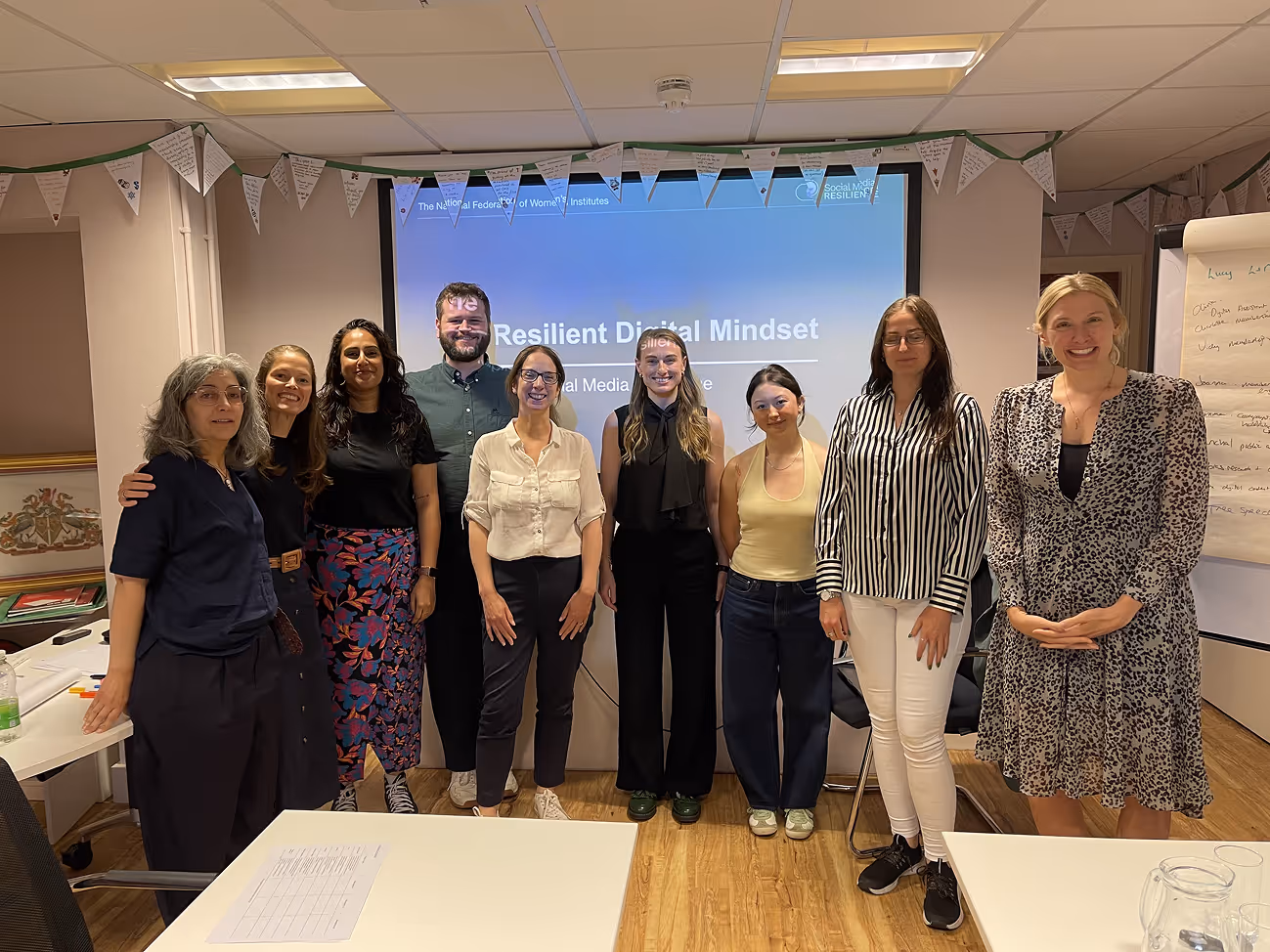 A group of smiling participants from the National Federation of Women's Institutes stand together at a Social Media Resilience "The Resilient Digital Mindset" stand in front of a screen. 