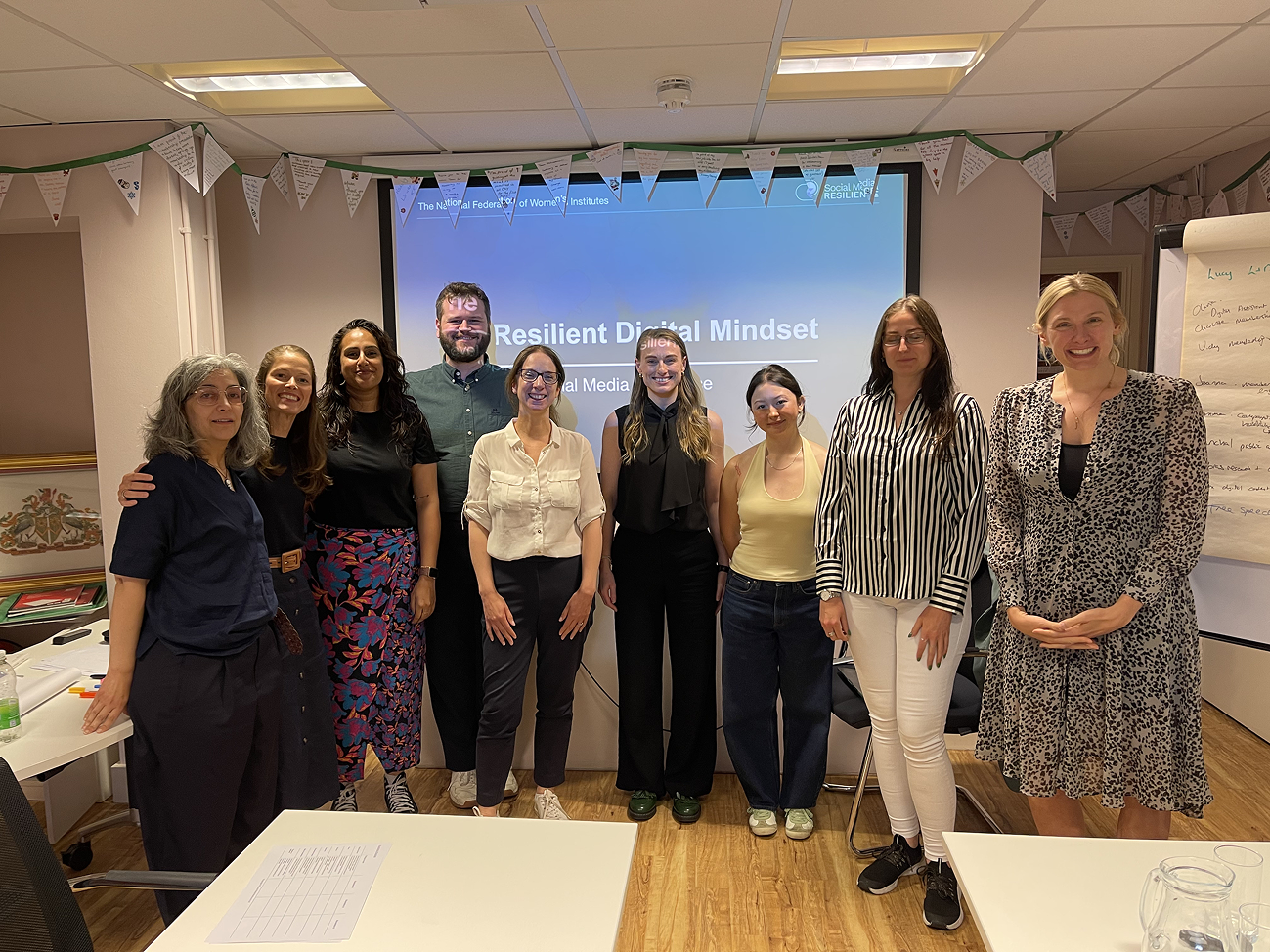 A group of smiling participants from the National Federation of Women's Institutes stand together at a Social Media Resilience "The Resilient Digital Mindset" stand in front of a screen. 