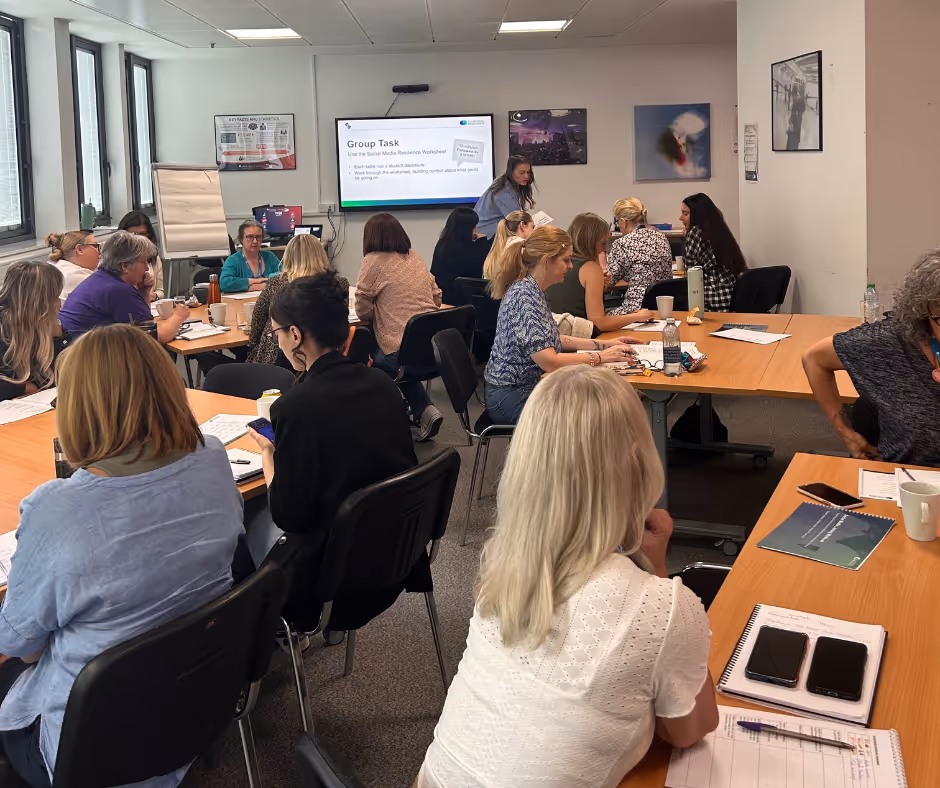 A group of participants in a Social Media Resilience Training session. there are workbooks on the tables, people looking towards the front of the room at a slide deck that says 'Group Task' with the trainer supporting participants. 
