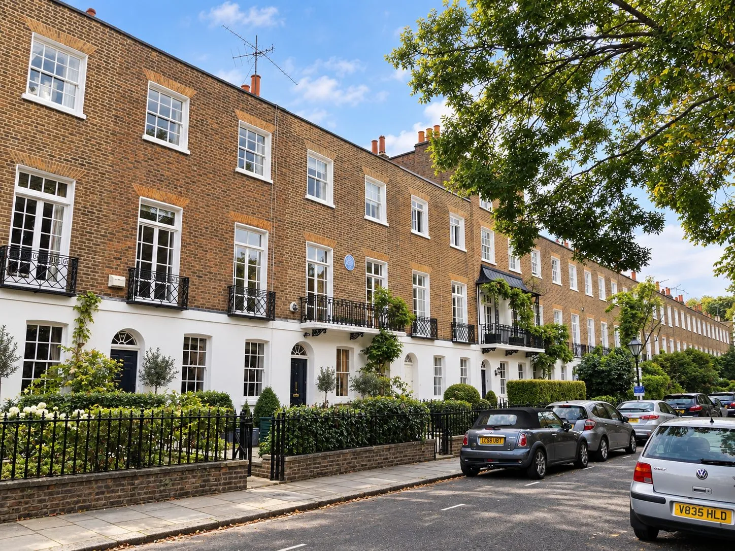 A terrace of Georgian brick houses with white stucco ground floors and iron balconies on a tree lined London street