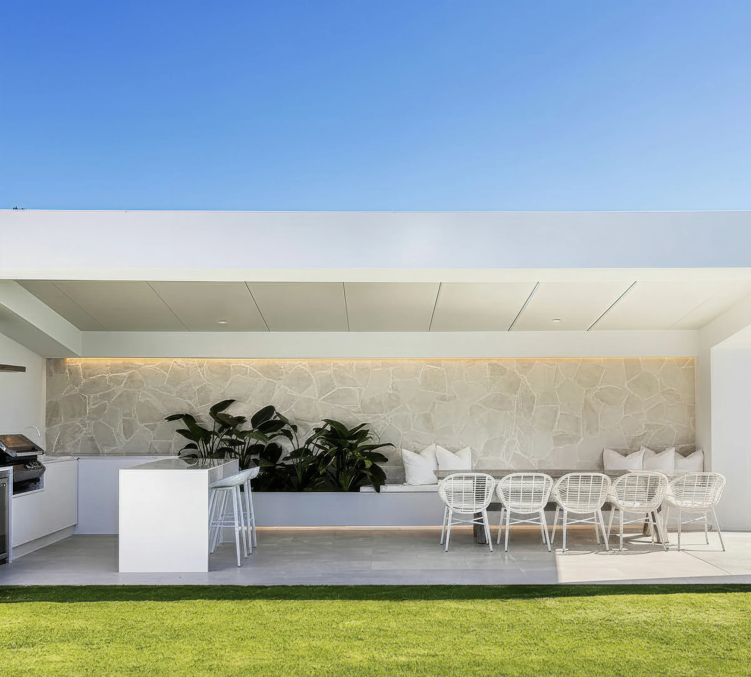 Covered outdoor venue with a stone wall, bar counter and white dining chairs beside a lawn