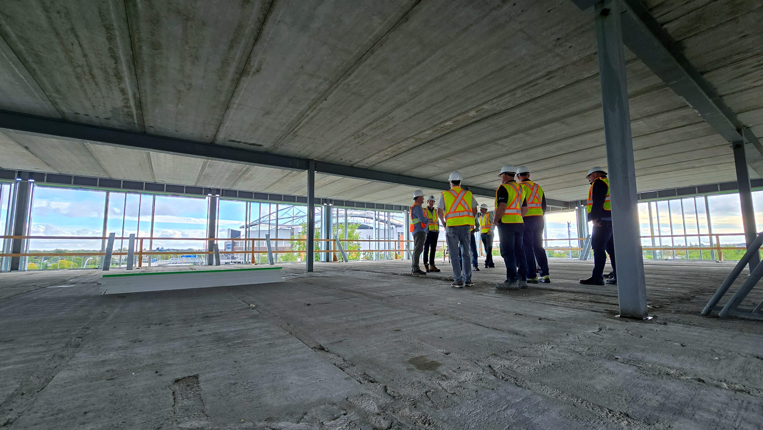 A group wearing yellow safety vests and hard hats stands on a concrete floor of a building under construction. The room is open with steel beams and large windows.