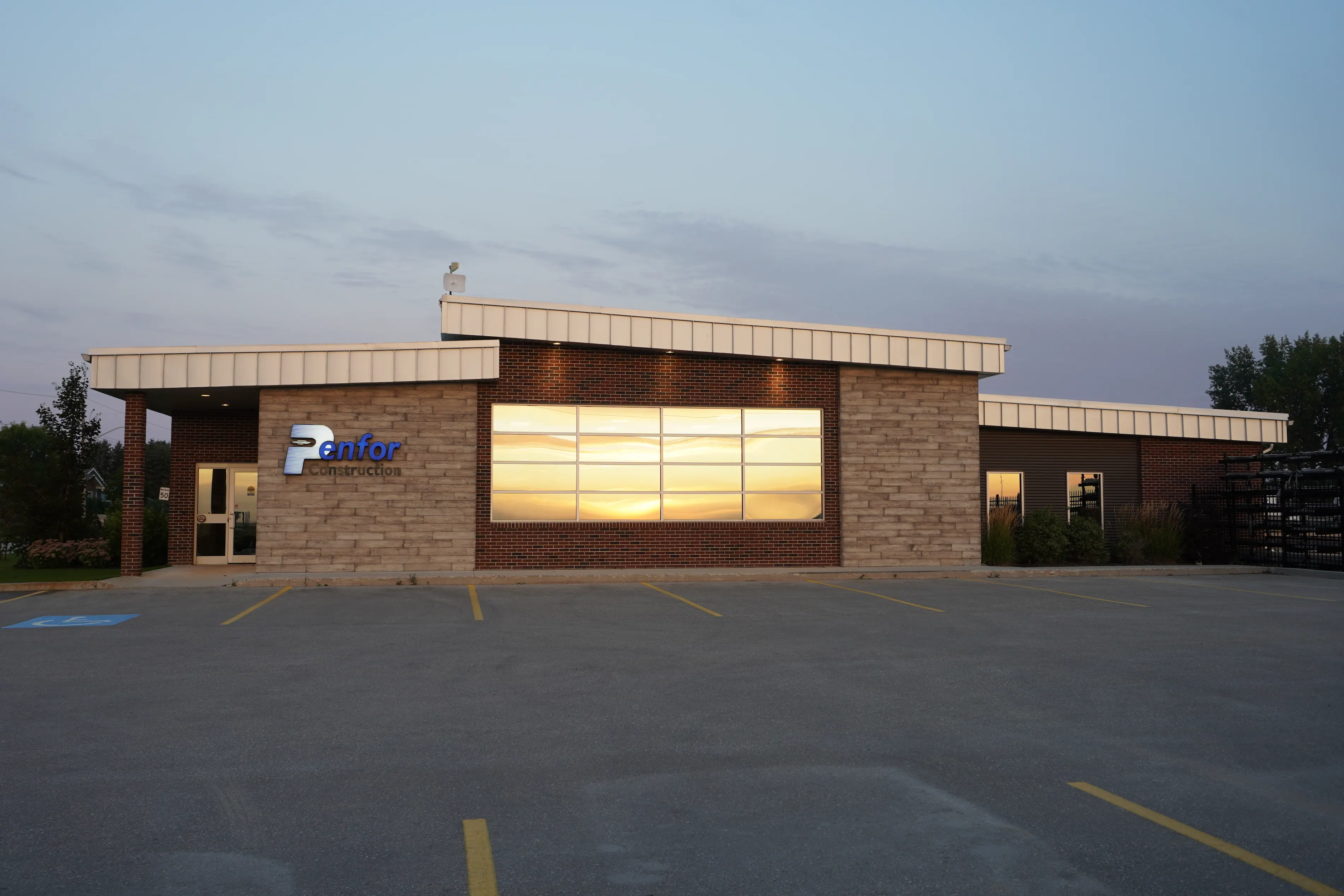 Single-story building at dusk with Penfor Construction signage and large windows reflecting a sunset. Made of stone and brick, with a calm atmosphere.