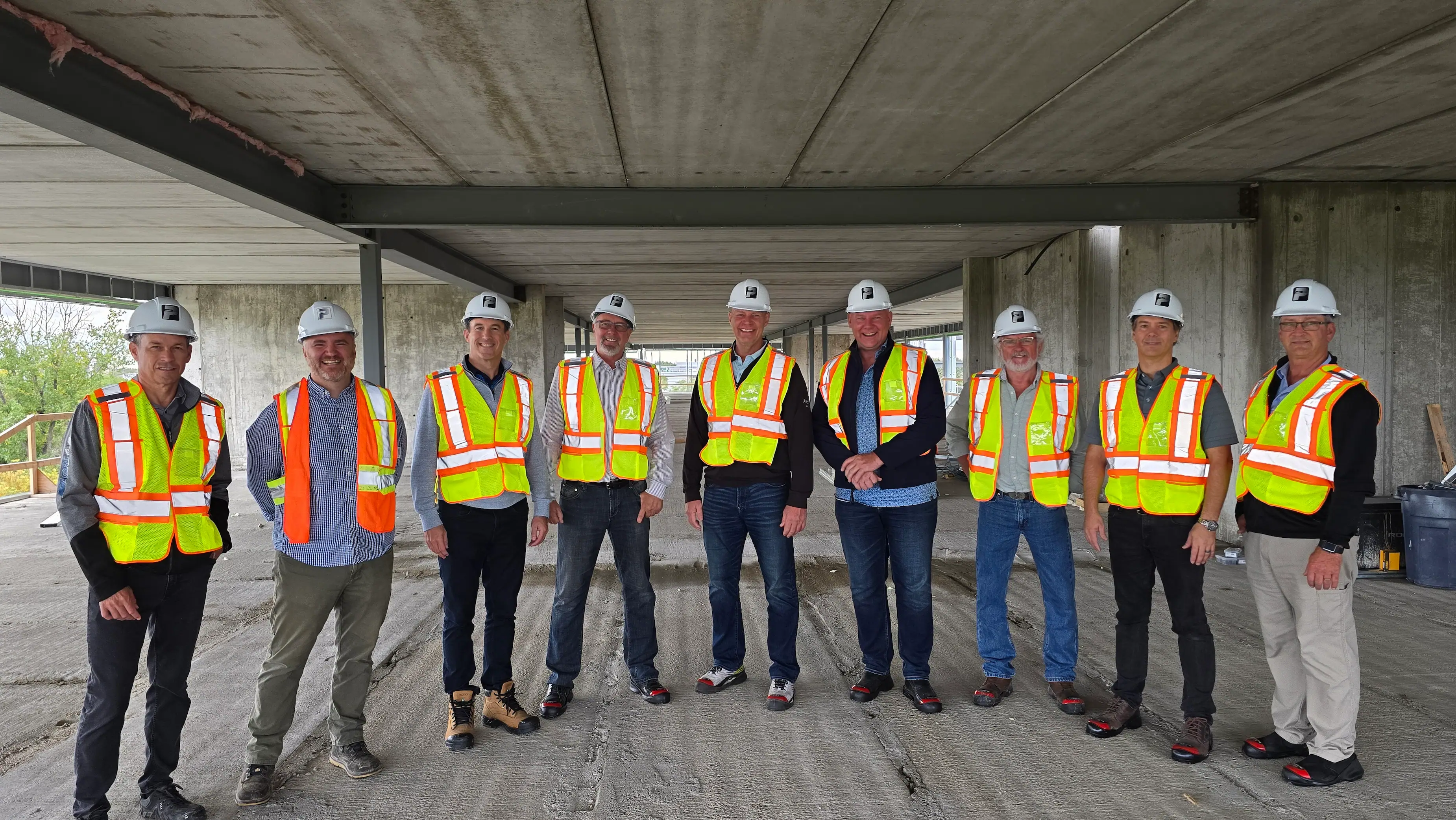 Nine people wearing hard hats and reflective safety vests stand in a line at a construction site.