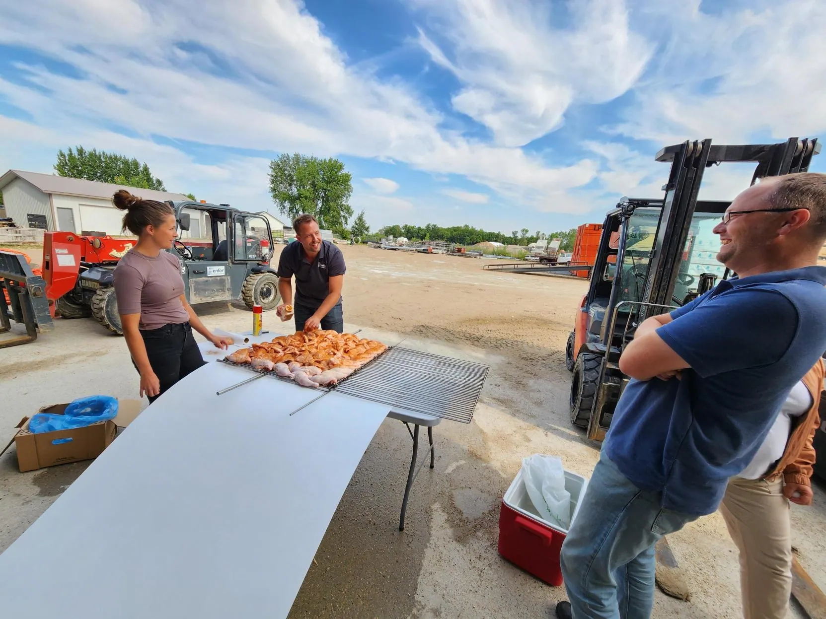 Three people are outside near a table with skewered meat, smiling and talking. Background includes forklifts and a clear, blue sky. Casual, outdoor setting.