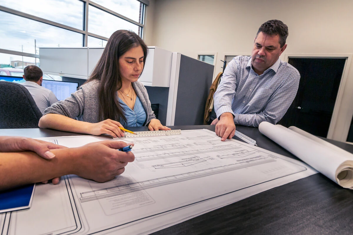A woman and two men review architectural blueprints in an office, focusing intently. The scene conveys collaboration and concentration.
