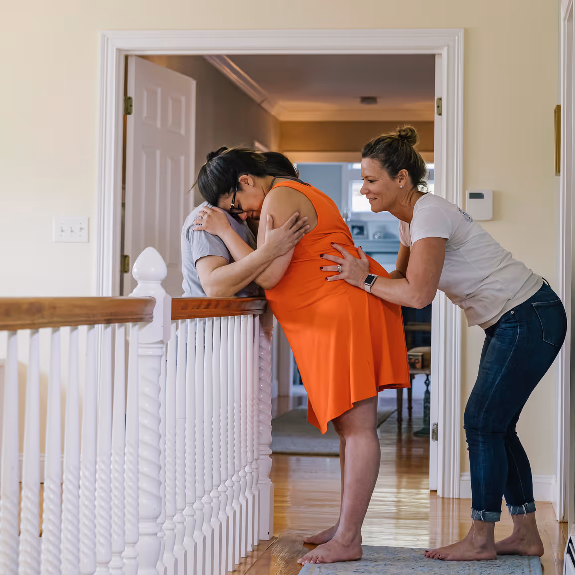 Doula giving a hip squeeze to a pregnant parent leaning onto their partner over a banister railing.