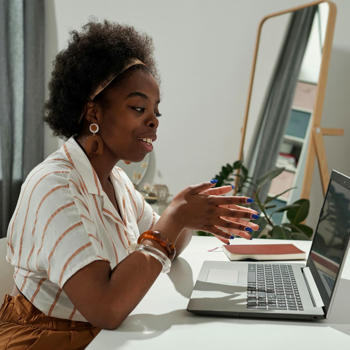 Black woman with natural hair working on laptop at home office desk, wearing striped blouse and blue nail polish