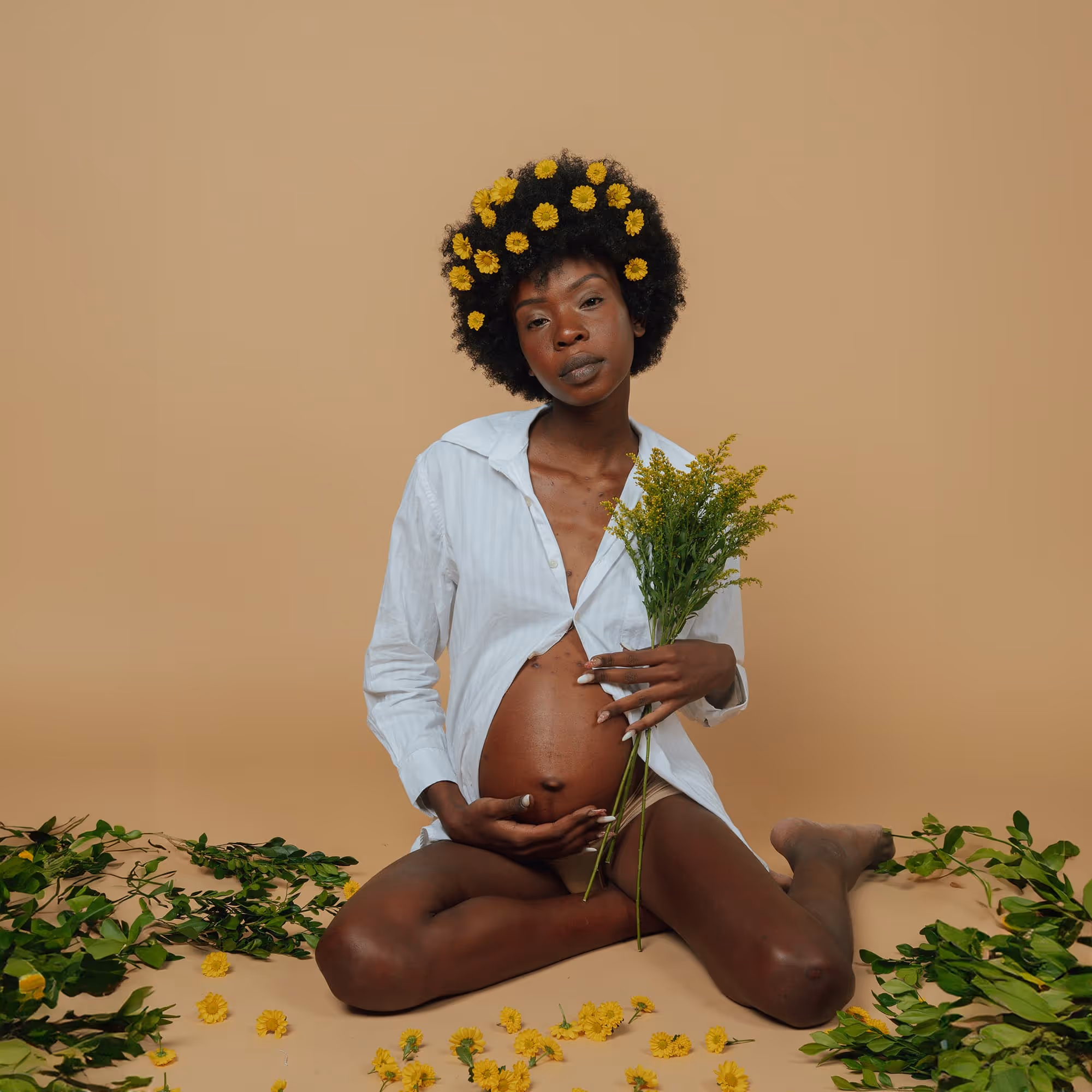 Black pregnant woman with yellow flowers in natural afro sitting cross-legged and holding belly in artistic maternity portrait