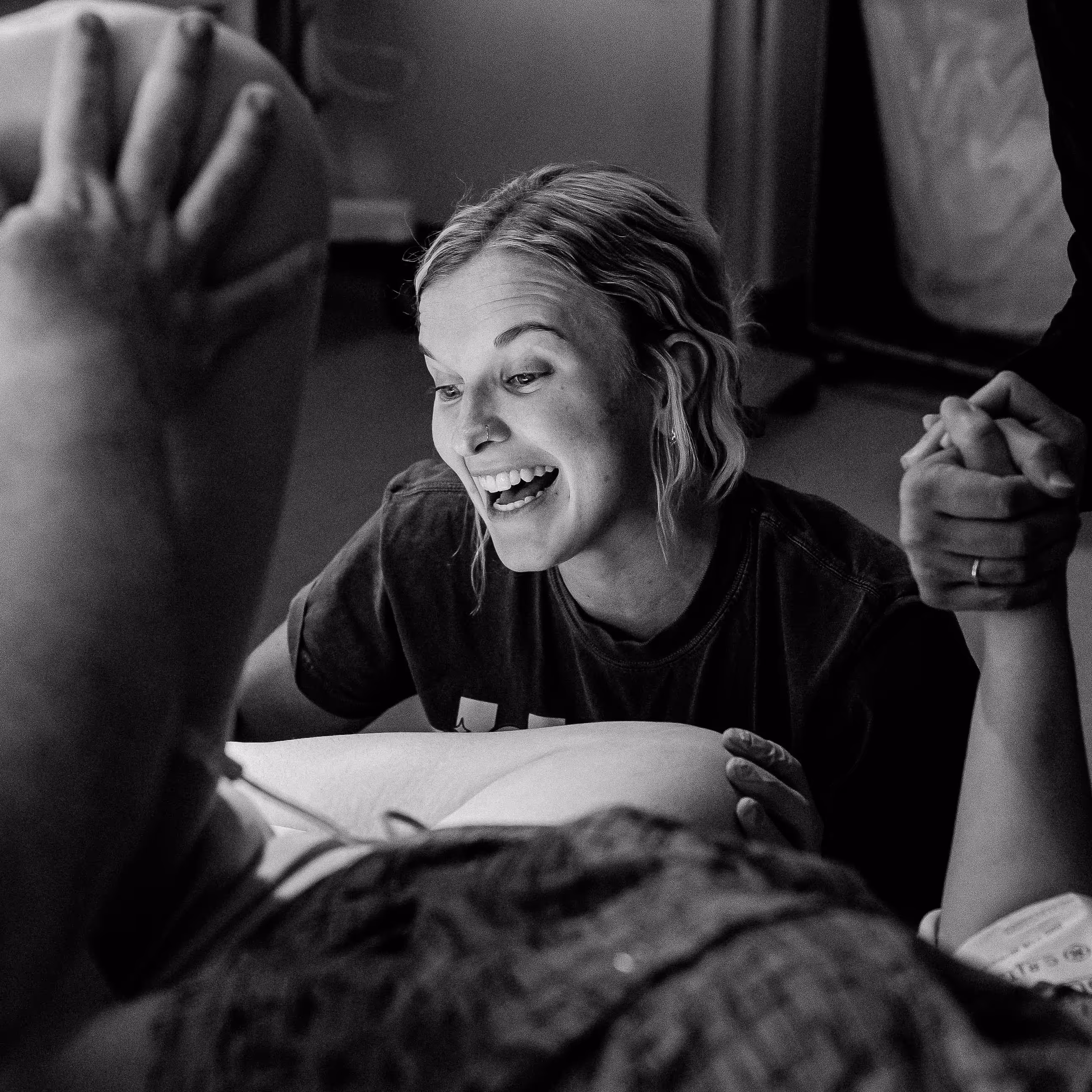 Black and white photograph of a smiling doula providing supportive care during labor, with her hands gently placed on a pregnant person's belly while a partner holds the laboring person's hand